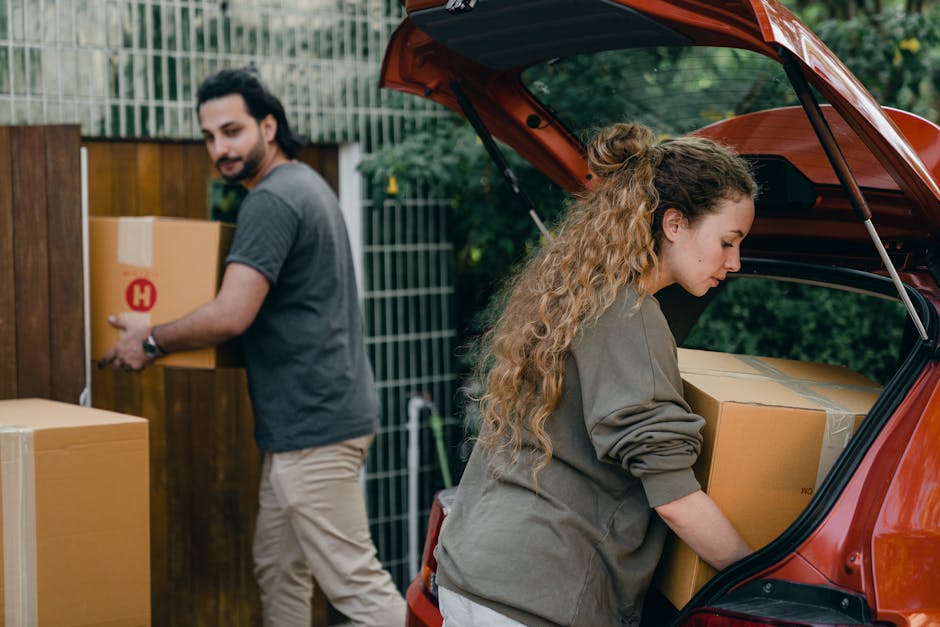 A man with long dark hair and a beard, dressed in a grey t-shirt and beige trousers, is transferring a large cardboard box with a red 'H' symbol in his hands, moving it toward a wooden fence outside a residential property. Nearby, a woman with long curly blonde hair, wearing a grey sweatshirt, is carefully placing a sizable cardboard box into the open trunk of a reddish-orange hatchback vehicle parked on the driveway. The vehicle's trunk is fully opened, exposing other packed boxes inside, and the scene is set in a garden with greenery and a wooden fence in the background. Several additional cardboard boxes sealed with packing tape are either on the ground or being loaded, illustrating a home relocation process involving packing and furniture transport, overseen by Man With a Van Kilburn as part of their removals services.