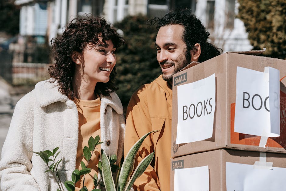 A man and a woman outdoors during a home relocation or moving day, standing beside a pile of cardboard boxes labeled 'BOOKS' and 'SMALL' in a residential area with trees and buildings in the background. The woman, wearing a cream-colored coat, is smiling at the man, who has dark curly hair and a beard, dressed in an orange jacket. The boxes are secured with packing tape, some wrapped in plastic, and there are houseplants visible in the foreground, indicating careful packing and preparation for a move. The scene takes place on a paved pavement or driveway, with natural daylight illuminating the subjects, capturing the loading process associated with furniture transport and packing during a house removal. As part of the services offered by Man With a Van Kilburn, such packing and loading activities are typical components of comprehensive moving logistics, ensuring safe and organized home relocation, aligning with the topic of Brent council move permits residents must know.