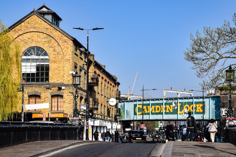 A street scene in Kilburn showing a brick building with large arched windows on the upper floor, adjacent to a railway bridge with a metal structure painted in green and yellow lettering spelling 'Camden Lock'. The scene includes parked cars, pedestrians walking along the pavement, and a tree with budding leaves on the right. Streetlights and a clock are mounted on poles along the road, with some construction cranes visible in the background under a clear blue sky. The image captures elements of urban environment consistent with home relocation and furniture transport, such as the traffic and pedestrian activity, which are relevant to house removals services offered by Man With a Van Kilburn in compliance with Camden council's waste and disposal guidelines.
