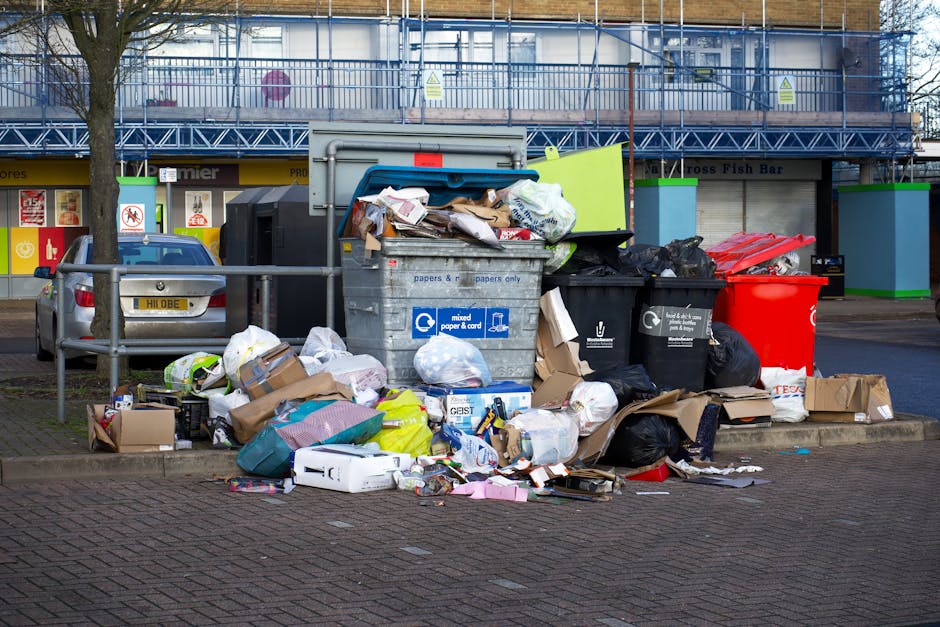 A large outdoor bin area near a commercial building in Kilburn, London, with several overflowing waste and recycling containers, including a grey mixed paper and card bin with an open lid, filled with flattened cardboard boxes, paper, and plastic waste. Surrounding the bins, there are numerous black and black plastic sacks, some torn open, revealing additional waste materials. Cardboard boxes and packaging are scattered on the pavement around the bins, alongside plastic bags and loose paper. A silver vehicle is parked adjacent to a waist-high metal barrier on the left side of the image. In the background, the building features signage for local shops including a fish bar, with a blue metal warehouse or scaffolding structure above, and trees with bare branches are visible on the left side of the scene. The setting appears to be during daytime in an urban area, illustrating waste management and disposal logistics relevant to house and property removals or home relocation services such as those provided by Man With a Van Kilburn.