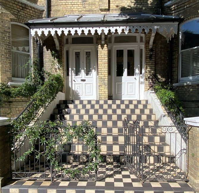 Image of the exterior entrance to a residential property during a house removal process by Man With a Van Kilburn, showing a staircase with black and beige checkered tiles leading up to two white front doors with decorative glass panels. The staircase is flanked by black wrought iron railings, with some greenery and bushes on either side. A collapsible metal gate is positioned at the bottom of the stairs, partially open, and a small metal ramp is placed on the left side, supporting the loading and unloading of furniture. The building has brick walls, with a covered porch area featuring a black canopy with decorative trim. This scene captures the careful arrangement during packing and furniture transport in a home relocation, highlighting the use of appropriate equipment for safe and efficient moving logistics.