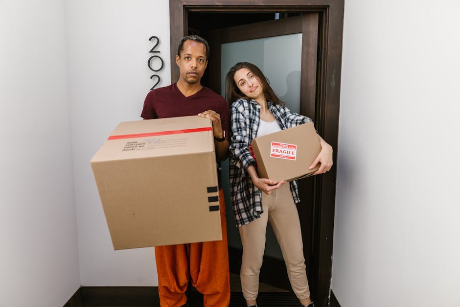A man with a serious expression and a woman with a slight smile are standing inside a building in front of a door with the number '200' visible on the wall beside it. The man is holding a large cardboard box with red tape and black markings, while the woman is holding a smaller cardboard box labeled 'Fragile' with red and white tape. Both are dressed casually; the man in a maroon T-shirt and orange trousers, and the woman in a plaid shirt and beige pants. The background shows a plain grey and white interior wall with the door frame made of dark wood. The scene captures a home relocation or furniture transport phase, and the environment is well-lit with natural or artificial lighting. This image illustrates packing and moving activities, with items prepared for transportation by Man With a Van Kilburn, reflecting the process of loading boxes in a house removal or moving service context.