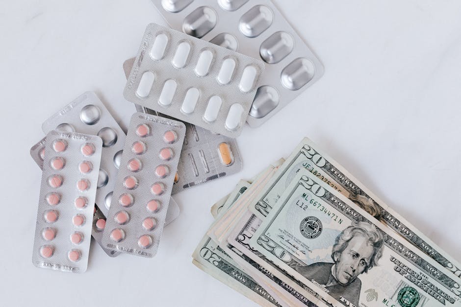 A collection of various blister packs of pills and tablets, including pink, white, and orange capsules, arranged next to a stack of US twenty-dollar bills on a plain white surface. The pills are in blister packaging made of clear plastic with aluminum backing, with some packs overlapping each other, and some pills missing from certain blisters. The twenty-dollar bills are folded and neatly stacked, positioned adjacent to the medication, suggesting an association with health-related expenses or costs. The image is well-lit, with a focus on the medication and money, reflecting themes of medical costs, pharmacy supplies, or health-related packaging in a professional context related to home relocation or moving logistics, as seen through the context of Man With a Van Kilburn's moving services.
