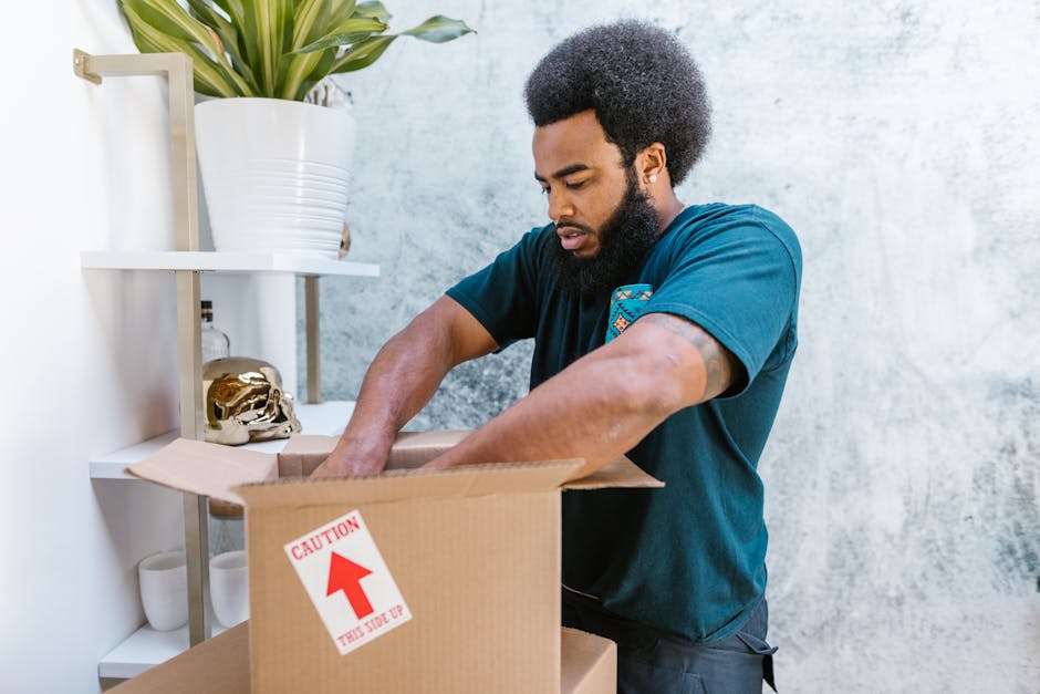 A man with a dark curly afro and beard, dressed in a navy blue t-shirt, is standing inside a home and packing items into an open cardboard box labeled with a red caution arrow and the words 'THIS SIDE UP.' He is reaching into the box with his right hand, carefully placing or adjusting belongings as part of a home relocation or packing process. In the background, there is a white wall with a shelf holding a potted plant with long green leaves, a small golden skull ornament, and white ceramic cups. The scene is well-lit with natural or soft indoor lighting, emphasizing the careful handling of items in preparation for moving or furniture transport, supported by professional removal services such as Man With a Van Kilburn.
