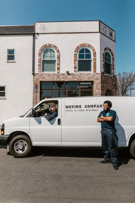A white moving van with the text 'MOVING COMPANY' and 'LOCAL & LONG DISTANCE' parked in front of a two-storey building with a brick and white façade. Inside the van, the driver is visible through the open window, wearing a cap and sunglasses, engaging in conversation with a colleague standing outside the vehicle. The colleague is a man with dark curly hair and a beard, dressed in a dark blue shirt and trousers, standing with arms crossed and smiling. The scene appears to be part of a home relocation process, with the van ready for furniture transport and packing and moving tasks. The building behind has large windows on the ground floor and arched windows above, suggesting a commercial or residential property in Kilburn, London. The overall environment is outdoors on a clear day, suitable for loading or unloading household belongings as part of a professional removals service. The image illustrates the planning and logistics involved in house removals, involving vehicle loading, client interaction, and professional staff coordination, typical of services like those offered by [COMPANY_NAME].