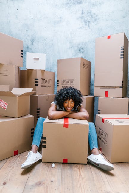 A young woman with curly black hair, wearing a white top and blue jeans, is sitting on the wooden floor inside a room surrounded by numerous cardboard boxes of various sizes, some sealed with red packing tape. She is smiling and resting her chin on a medium-sized box in front of her. The boxes are stacked along the walls, which have a textured light blue and white paint finish, indicating an interior space prepared for moving or packing. The scene captures the packing process involved in home relocation or furniture transport, which is associated with professional removal services such as those provided by Man With a Van Kilburn. The overall environment emphasizes organization and readiness for logistics involved in packing and moving furniture or household items.