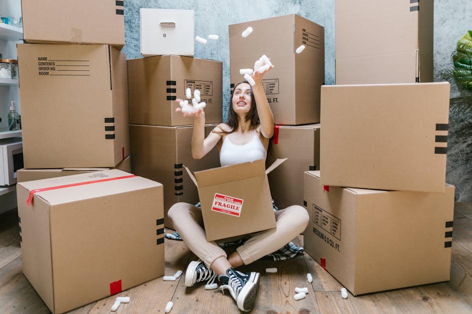 A young woman sitting cross-legged on a wooden floor surrounded by large cardboard moving boxes, some marked with 'Fragile' labels. She is wearing a white tank top, beige pants, and black sneakers with white laces. She is smiling and throwing packing foam peanuts into the air, with several foam pieces falling around her. The boxes are stacked in the background, some of which are inside an interior room with light-colored walls and a decorative plant visible on the right side. The scene depicts the packing and loading process during a home relocation, with the woman engaged in packing or preparing boxes for搬迁, representing services offered by Man With a Van Kilburn for furniture transport and removals, aligned with tips for accessing flats on Kilburn High Road.