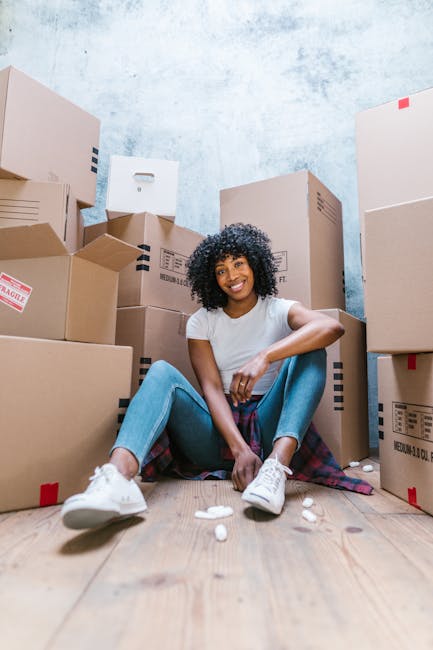 A young woman with curly black hair, wearing a white top and blue jeans, is sitting on the wooden floor inside a room surrounded by numerous cardboard boxes of various sizes, some sealed with red packing tape. She is smiling and resting her chin on a medium-sized box in front of her. The boxes are stacked along the walls, which have a textured light blue and white paint finish, indicating an interior space prepared for moving or packing. The scene captures the packing process involved in home relocation or furniture transport, which is associated with professional removal services such as those provided by Man With a Van Kilburn. The overall environment emphasizes organization and readiness for logistics involved in packing and moving furniture or household items.