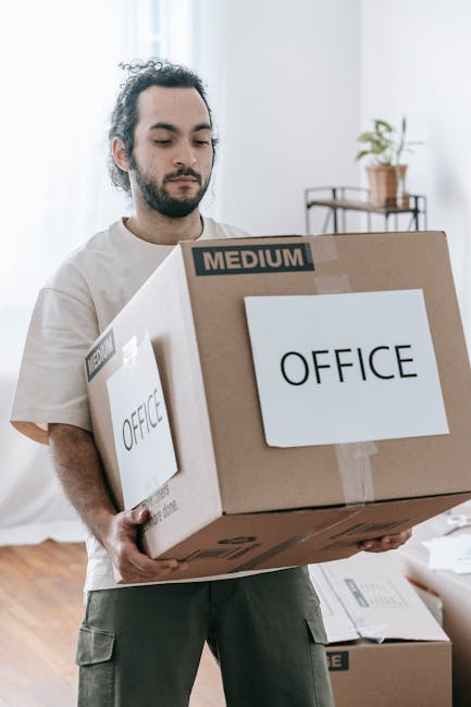 A man with dark, curly hair and a beard, wearing a light-colored t-shirt, is carefully carrying a medium-sized cardboard box labeled 'OFFICE' across a room with wooden flooring. The box is wrapped with clear packing tape and rests securely in his arms, with another similar box visible on the floor nearby. The background includes a doorway leading to a bright room with a small table holding a potted plant, reflecting a residential or office environment during a home or office relocation process. This scene depicts the packing and furniture transport stages of a house or business removal service, performed by Man With a Van Kilburn as part of their local removals offerings for Kilburn residents and businesses on High Road.