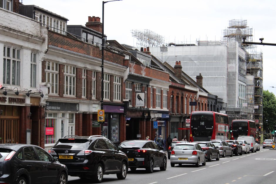 A street scene in Kilburn showing a line of parked cars and two red double-decker buses along High Road, with pedestrians walking on the pavement. Behind the vehicles, there are multiple historic brick buildings with white window frames, some commercial establishments including a shop with a red sign, and scaffolding covering a building under renovation. Inside the street, a moving company, Man With a Van Kilburn, is involved in a home relocation or furniture transport process, with visible cardboard boxes, wrapped furniture, and loading equipment such as trolleys or blankets possibly seen nearby as part of the moving operation. The scene is captured during daylight with overcast sky, highlighting the typical urban environment of Kilburn, where packing and moving activities take place in preparation for house or office removals, supporting relocation logistics.