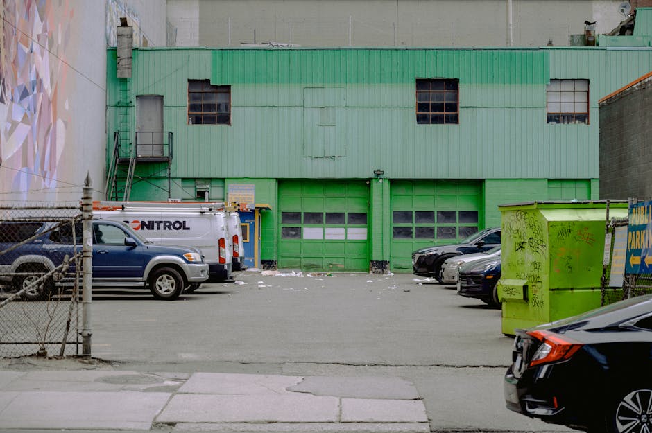 A green industrial building with three large garage doors and three small, square windows above them, situated in an urban setting. In front of the building, a paved parking area houses several vehicles, including a white van labelled 'NITROL' parked on the left side, and multiple passenger cars in various colors parked along the right. The area is enclosed by a chain-link fence on the left, with a small gate. To the right, a bright green refuse bin with graffiti and a yellow warning sign is positioned near the curb. The loading bay, located at the center of the building, is being used for home relocation activities, with available space for loading and unloading furniture, boxes, and packing materials. The scene depicts a typical environment for furniture transport and moving logistics, aligned with the rules for removal vans at Kilburn Park station, as managed by Man With a Van Kilburn.