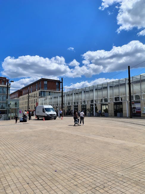 A green industrial building with three large garage doors and three small, square windows above them, situated in an urban setting. In front of the building, a paved parking area houses several vehicles, including a white van labelled 'NITROL' parked on the left side, and multiple passenger cars in various colors parked along the right. The area is enclosed by a chain-link fence on the left, with a small gate. To the right, a bright green refuse bin with graffiti and a yellow warning sign is positioned near the curb. The loading bay, located at the center of the building, is being used for home relocation activities, with available space for loading and unloading furniture, boxes, and packing materials. The scene depicts a typical environment for furniture transport and moving logistics, aligned with the rules for removal vans at Kilburn Park station, as managed by Man With a Van Kilburn.