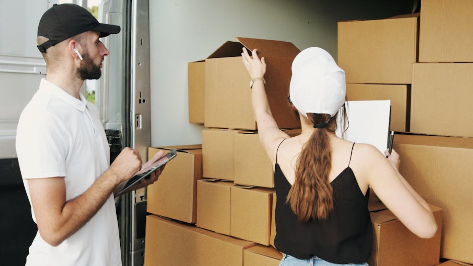 A male removal worker wearing a black cap, white t-shirt, and wireless earbud is standing next to an open box truck, holding a clipboard and pen, while a female worker with long brown hair, wearing a 