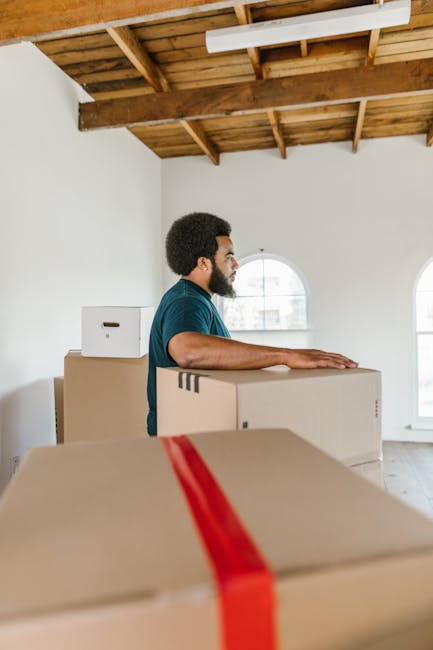A man with dark curly hair and a beard, wearing a blue T-shirt, is indoors in a spacious room with white walls, a wooden ceiling with exposed beams, and two arched windows allowing natural light. He is standing behind a stack of cardboard moving boxes, some with visible packing tape, and appears to be preparing for home relocation or furniture transport. Additional boxes are placed on the floor and on top of others nearby. The scene captures the loading process as part of packing and moving activities, with no furniture or large appliances visible. The image is associated with residential moving services, possibly provided by Man With a Van Kilburn, and depicts a typical packing and loading stage during a house removal or end-of-term move in Kilburn.