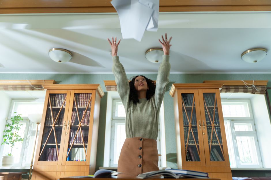 A woman with curly dark hair is standing inside a room with light green walls and large windows, lifting a sheet of paper or cardboard towards the ceiling. She is wearing a light grey ribbed sweater and a tan skirt with buttons down the front. Behind her, there are two wooden display cabinets filled with books, positioned on either side of the window and partially blocking natural daylight. A potted plant is visible on a windowsill to the left. The ceiling has three round, flush-mounted light fixtures, and the room appears well-lit. The scene captures a moment during packing or unpacking as part of a home relocation or moving process, with the woman possibly organizing or preparing items for transportation, which is relevant to furniture transport and packaging services often provided by companies like Man With a Van Kilburn.