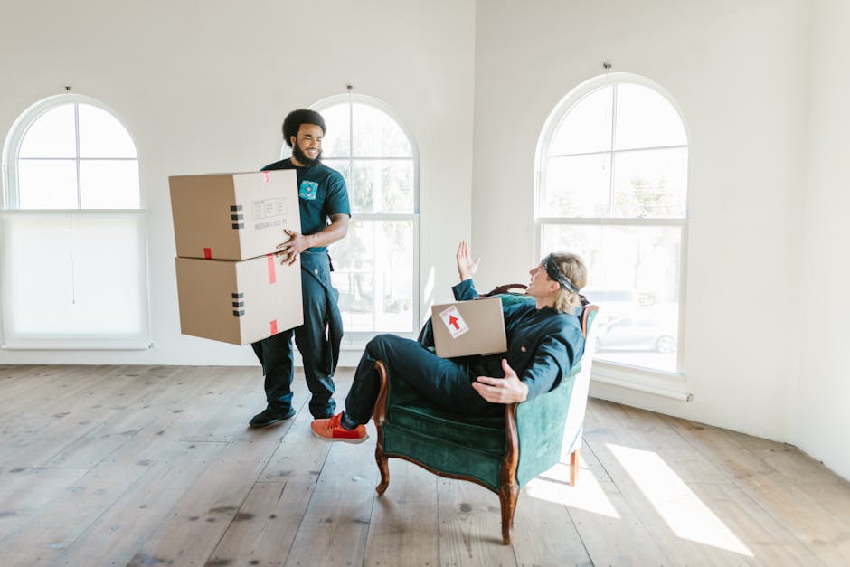 A man with dark curly hair and a beard, wearing a blue T-shirt, is indoors in a spacious room with white walls, a wooden ceiling with exposed beams, and two arched windows allowing natural light. He is standing behind a stack of cardboard moving boxes, some with visible packing tape, and appears to be preparing for home relocation or furniture transport. Additional boxes are placed on the floor and on top of others nearby. The scene captures the loading process as part of packing and moving activities, with no furniture or large appliances visible. The image is associated with residential moving services, possibly provided by Man With a Van Kilburn, and depicts a typical packing and loading stage during a house removal or end-of-term move in Kilburn.
