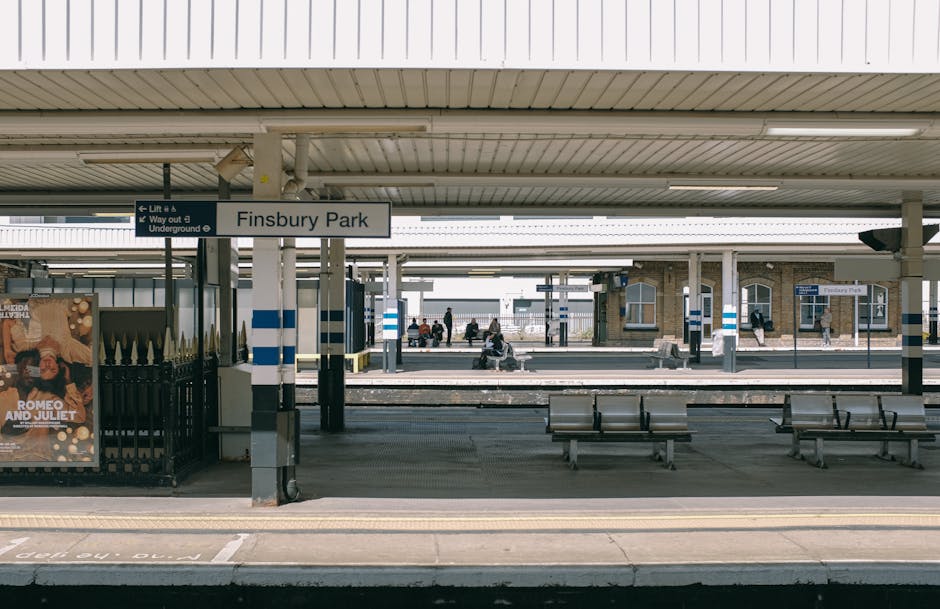 The image depicts Finsbury Park train station platform in daytime, with a large white sign displaying the station name. The platform is empty in the foreground, with several metal benches positioned along the paved area. Overhead, the station's roof extends across the platform, supported by evenly spaced columns painted with blue and white stripes. In the background, a few passengers are seated on benches or walking, while others are waiting near the train tracks. To the left, there is a black metal fence with a poster advertising a production of 'Romeo and Juliet.' The station environment appears clean, organized, and well-lit by natural daylight, illustrating a typical setting for home relocation or moving logistics involving train travel, with visible platforms, waiting areas, and signage that Man With a Van Kilburn might coordinate with for efficient furniture transport or packing and moving services.