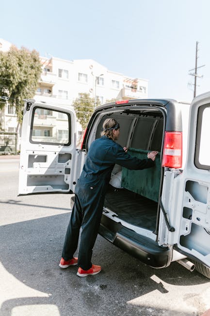 A person dressed in dark clothing and red shoes is standing beside an open white cargo van parked on a paved street, engaged in the loading process. They are placing or adjusting a large, rectangular green item inside the van, which is lined with a black interior. The van’s rear doors are fully open, revealing the spacious cargo area with minimal items inside. The scene takes place outdoors under bright daylight, with residential buildings, trees, and a utility pole visible in the background. The environment and positioning suggest an active home relocation or furniture transport operation, with the individual preparing items for a house moving service provided by Man With a Van Kilburn, supporting efficient packing and logistical planning for a same-day move in Kilburn NW6.