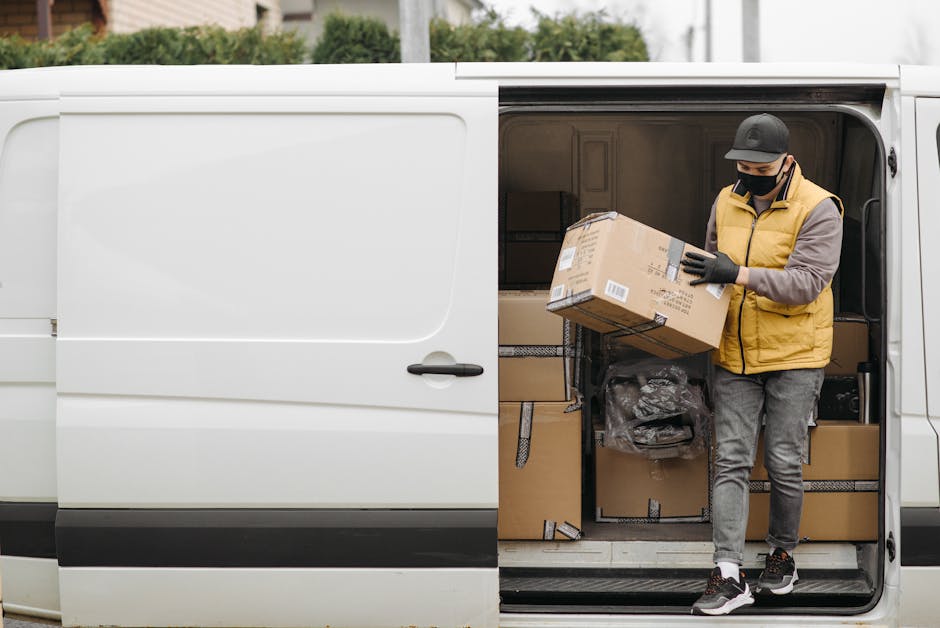 A man wearing a black cap, face mask, beige puffer vest, grey pants, and black sneakers is standing inside the open side door of a white delivery van. He is holding a cardboard box with printed labels and barcodes, preparing to load or unload it. The van's interior is filled with multiple packed cardboard boxes, some sealed with black tape, and assorted packing materials. The van is parked on pavement, surrounded by a blurred outdoor background with trees and greenery. The scene depicts the logistics of packing and home relocation, with the man actively engaged in furniture transport or moving services, coordinated by Man With a Van Kilburn as part of their removals process.